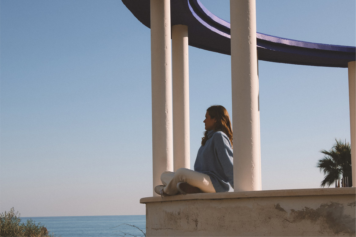 Merce sitting calmly in a white-columned pavilion above the sea